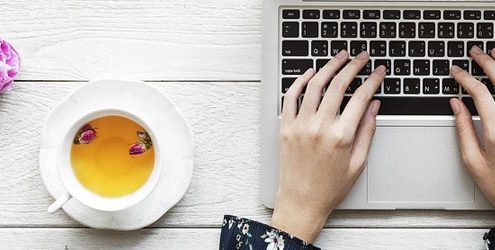 Mani di donna che scrivono su un notebbok appoggiato su un tavolo di legno bianco insieme a dei fiori e una tazzina di caffè