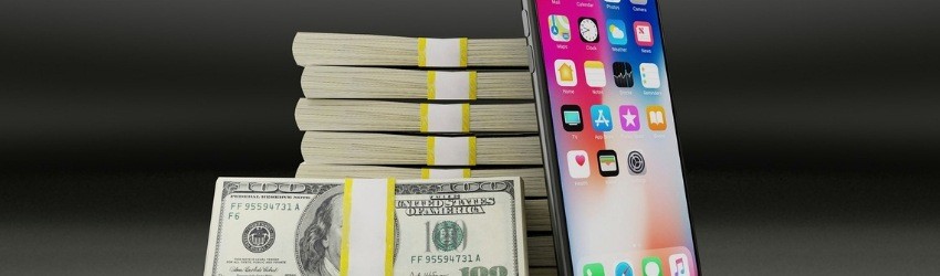 pila di banconote rilegate con una fascetta a cui sono appoggiati una fascetta di 100 dollari e un I-Phone acceso su sfondo grigio scuro