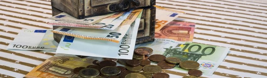 piccola cassaforte da cui fuoriescono banconote euro sopra ad un tavolo dove sono sparpagliate altre banconote e monete euro
