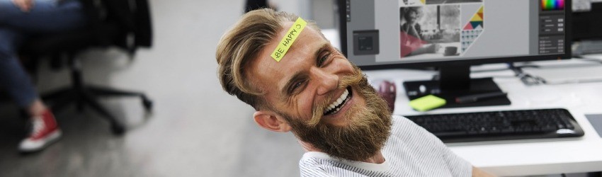 Uomo sorridente davanti a un computer con post-it giallo in fronte con scritto "be happy" con