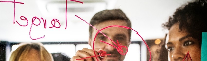 Pannello trasparente con schema rosso target su sfondo con un uomo che disegna lo schema e due donne che lo guardano